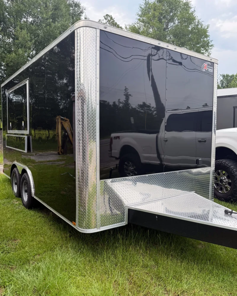 Black enclosed dual-axle cargo trailer with diamond plate trim, parked on grass beside a white pickup truck